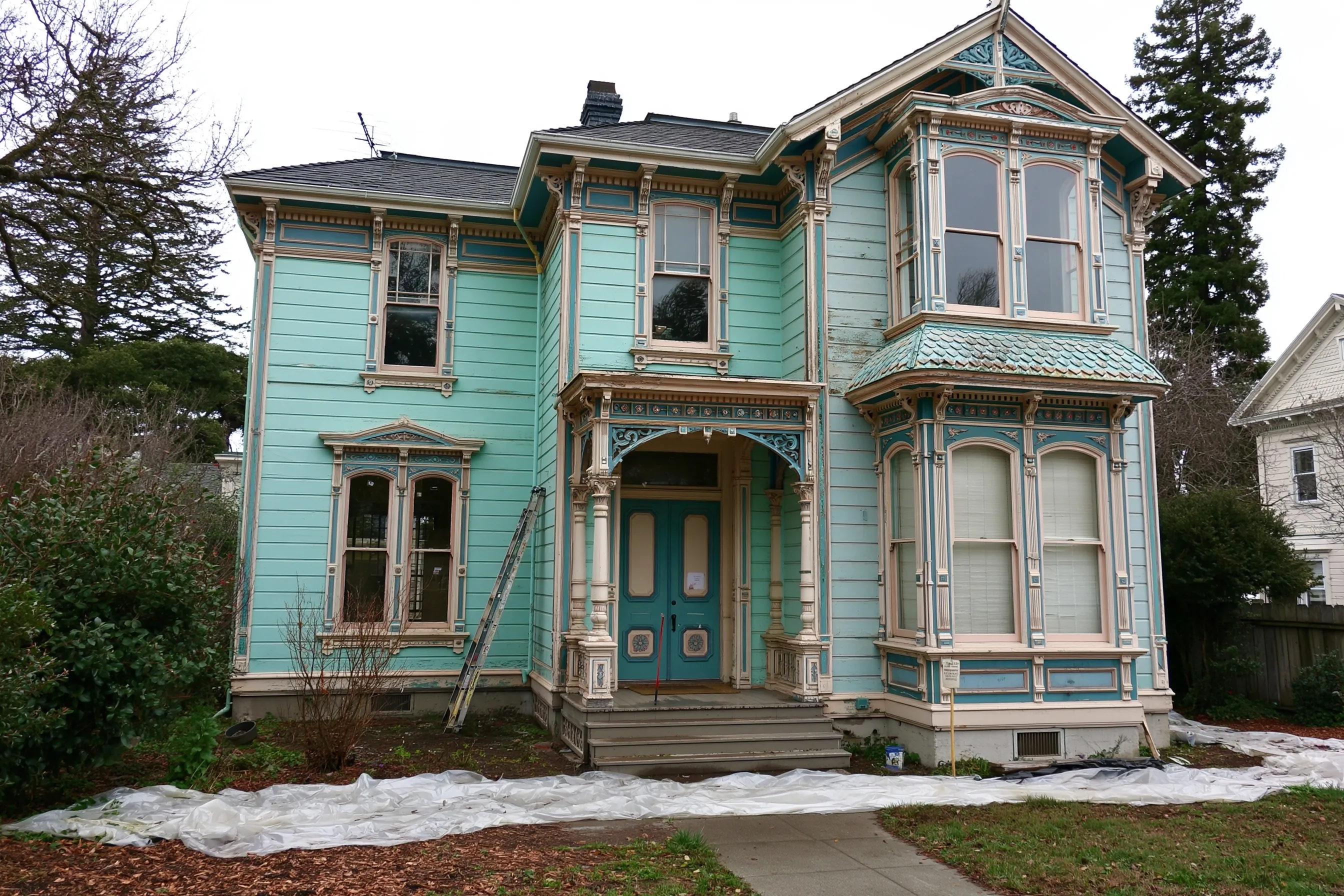 Two-story Victorian house with painted wood siding and ornate trim, a ladder against the wall and plastic sheeting on the ground in front